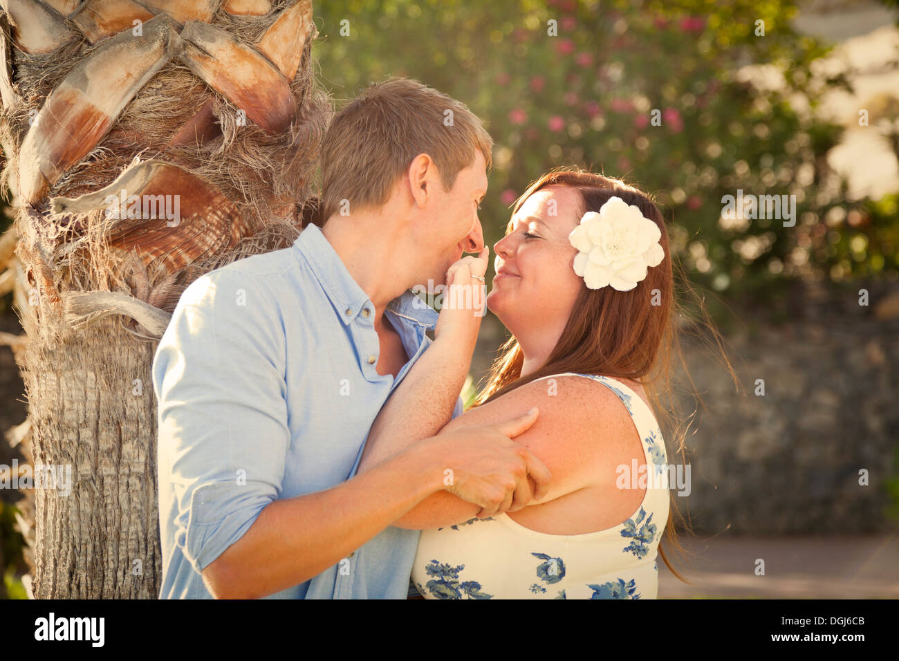 Playful couple kissing and teasing in sub tropical garden with palm trees and flowers, in late evening warm, soft light. Stock Photo