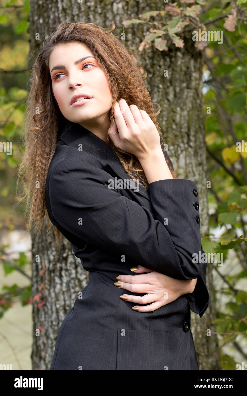 Young woman with long hair wearing a black blazer in front of a tree Stock Photo
