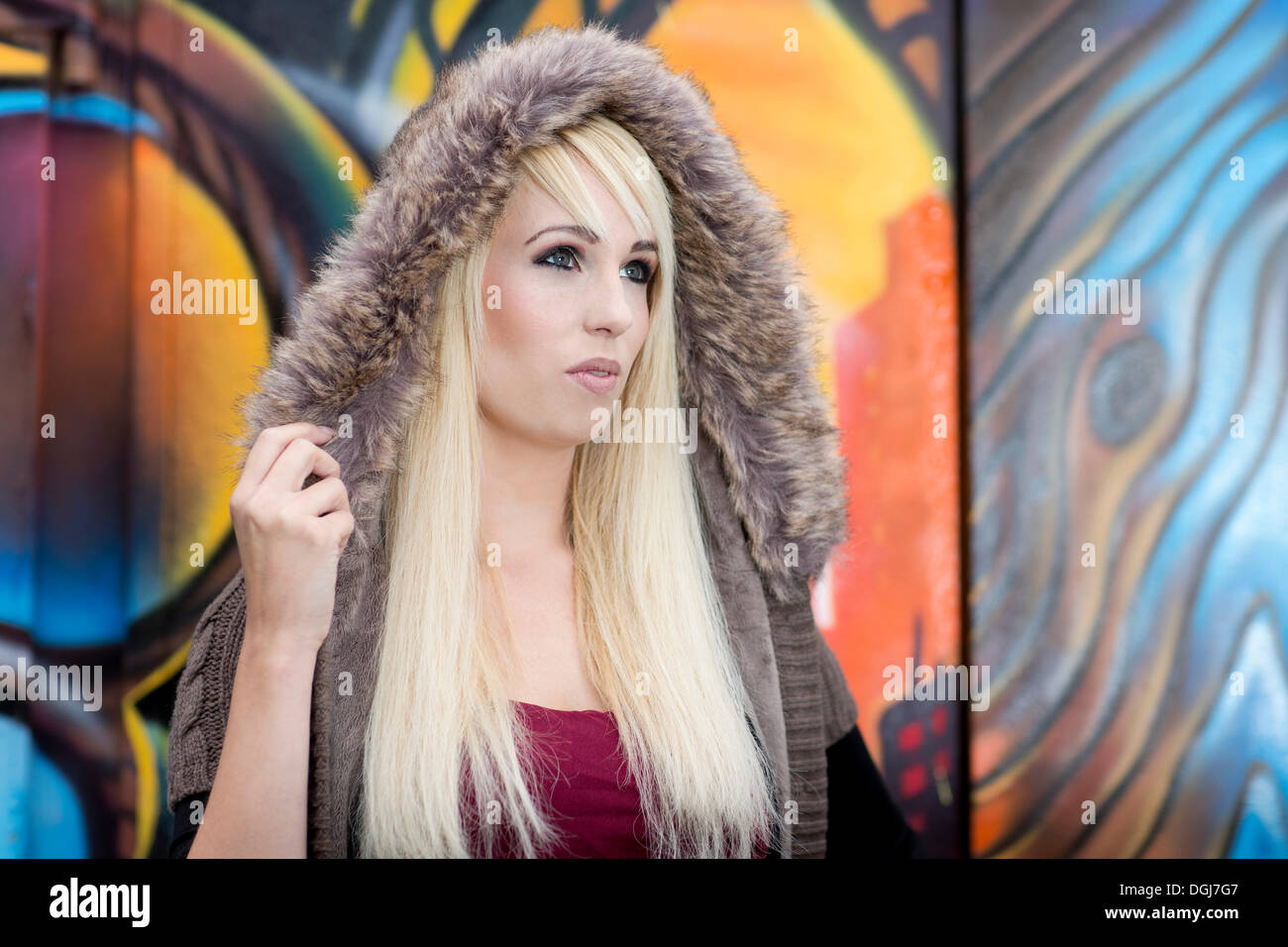 Young woman with long blonde hair wearing a fur hood posing in front of a wall with graffiti Stock Photo