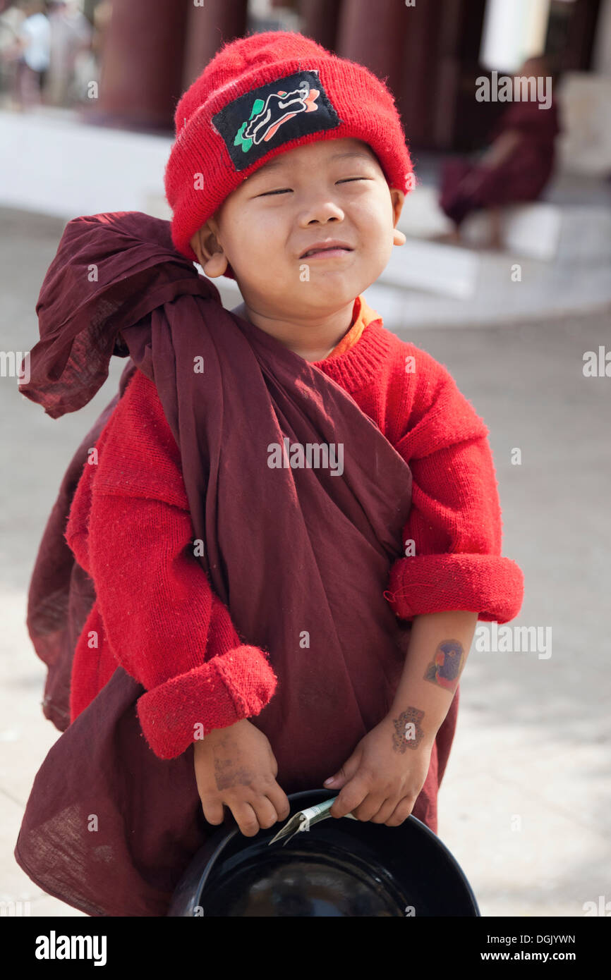 A tiny boy in Monk's robes and red hat with a his collection bowl in Shwezigon Pagoda in Bagan in Myanmar. Stock Photo