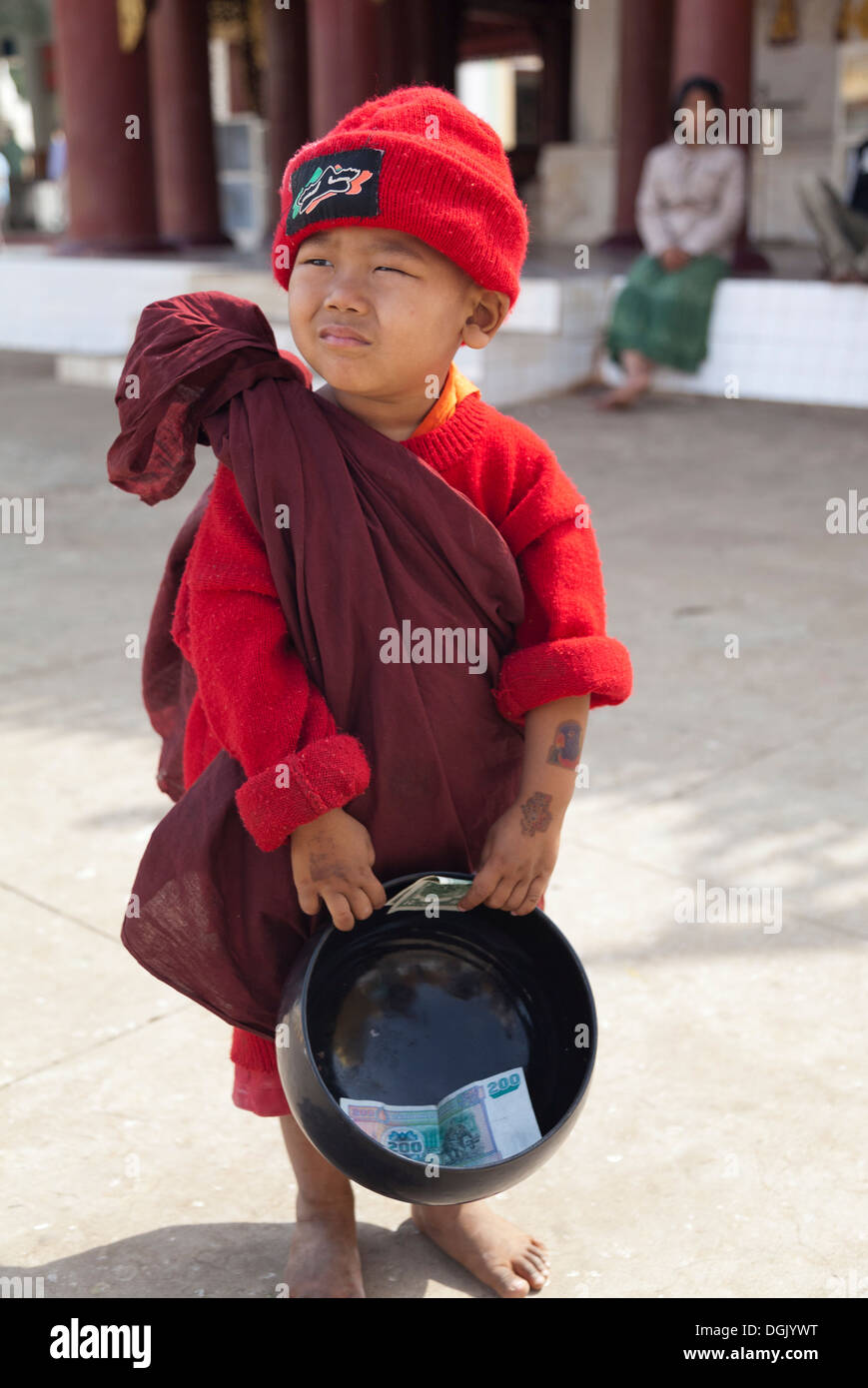 A tiny boy in Monk's robes and red hat with a his collection bowl in Shwezigon Pagoda in Bagan in Myanmar. Stock Photo