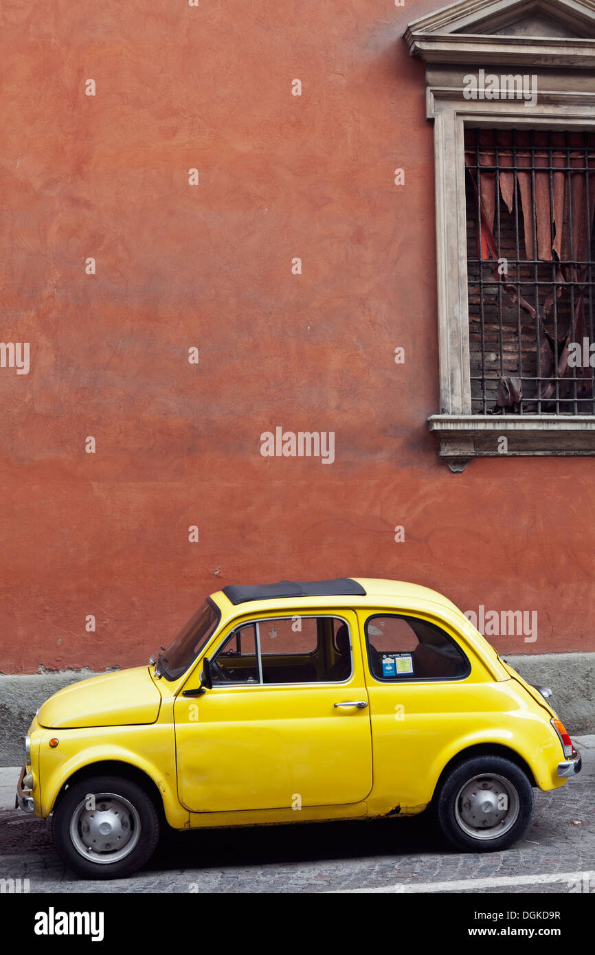 A vintage yellow Fiat 500 car parked on a cobbled street in front of an ochre wall in Bologna. Stock Photo