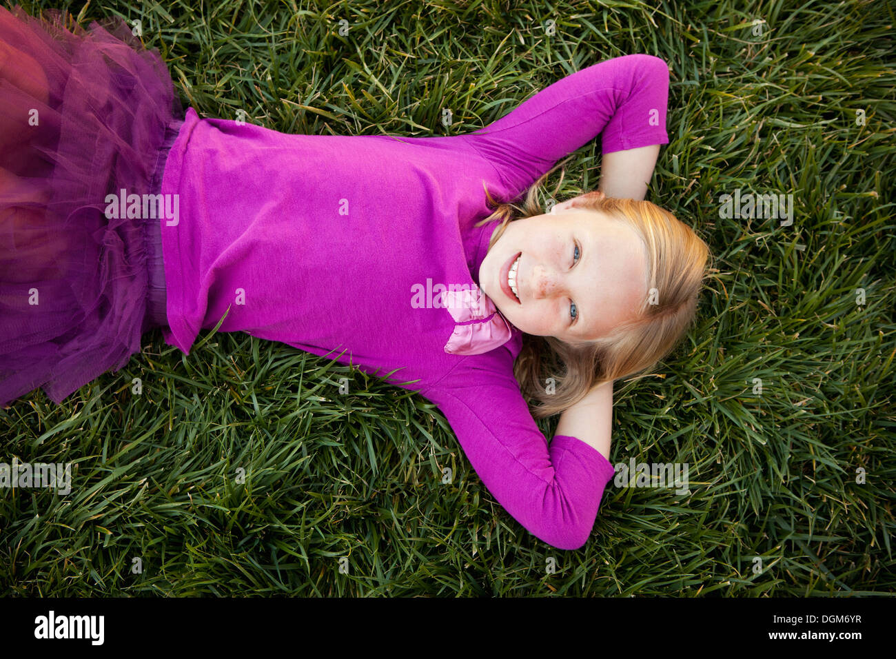 A young girl lying on her back on grass her hands behind her head smiling looking camera View from above Stock Photo