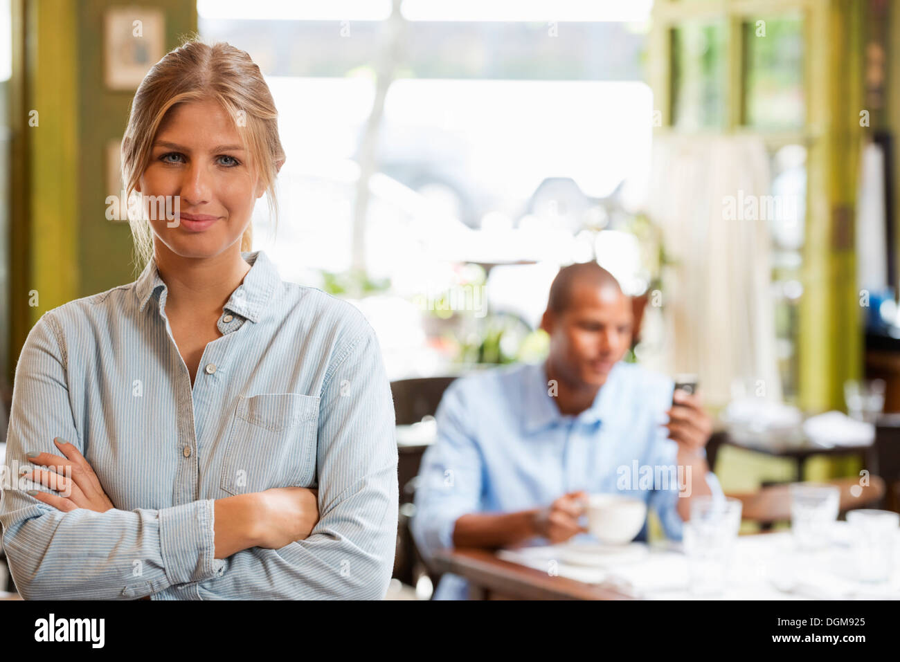 A couple in a city coffee shop. A woman sitting down checking a smart phone. A man standing up with arms folded. Stock Photo