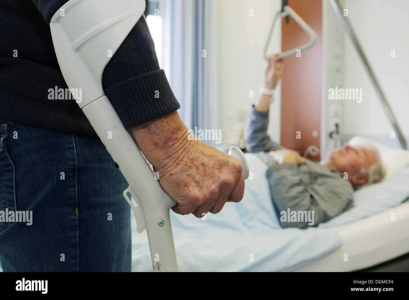 Elderly woman standing on a crutch in front of an elderly man lying in a hospital bed Stock Photo