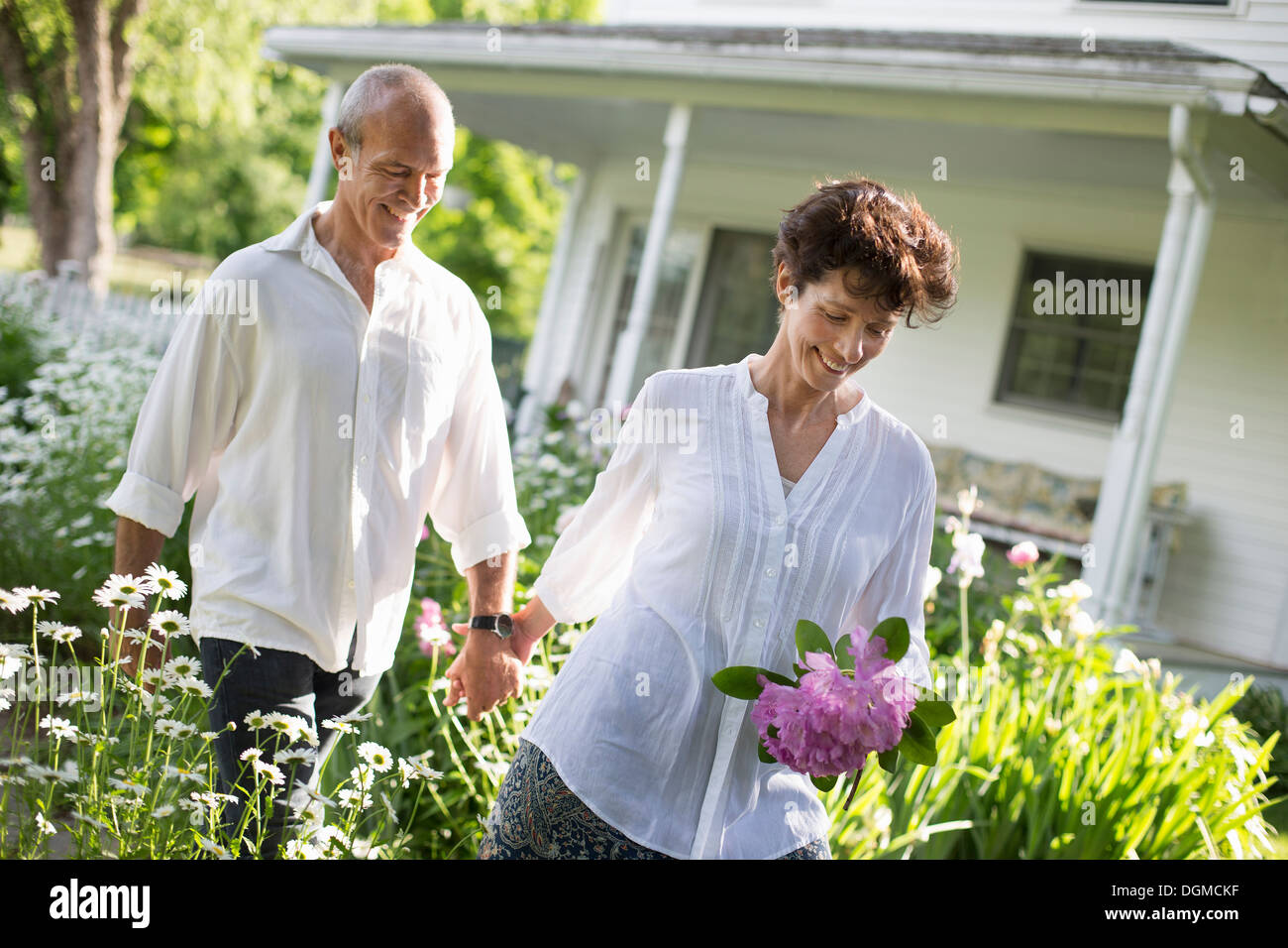 Organic farm. Summer party. A mature couple in white shirts walking holding hands through the garden. Stock Photo