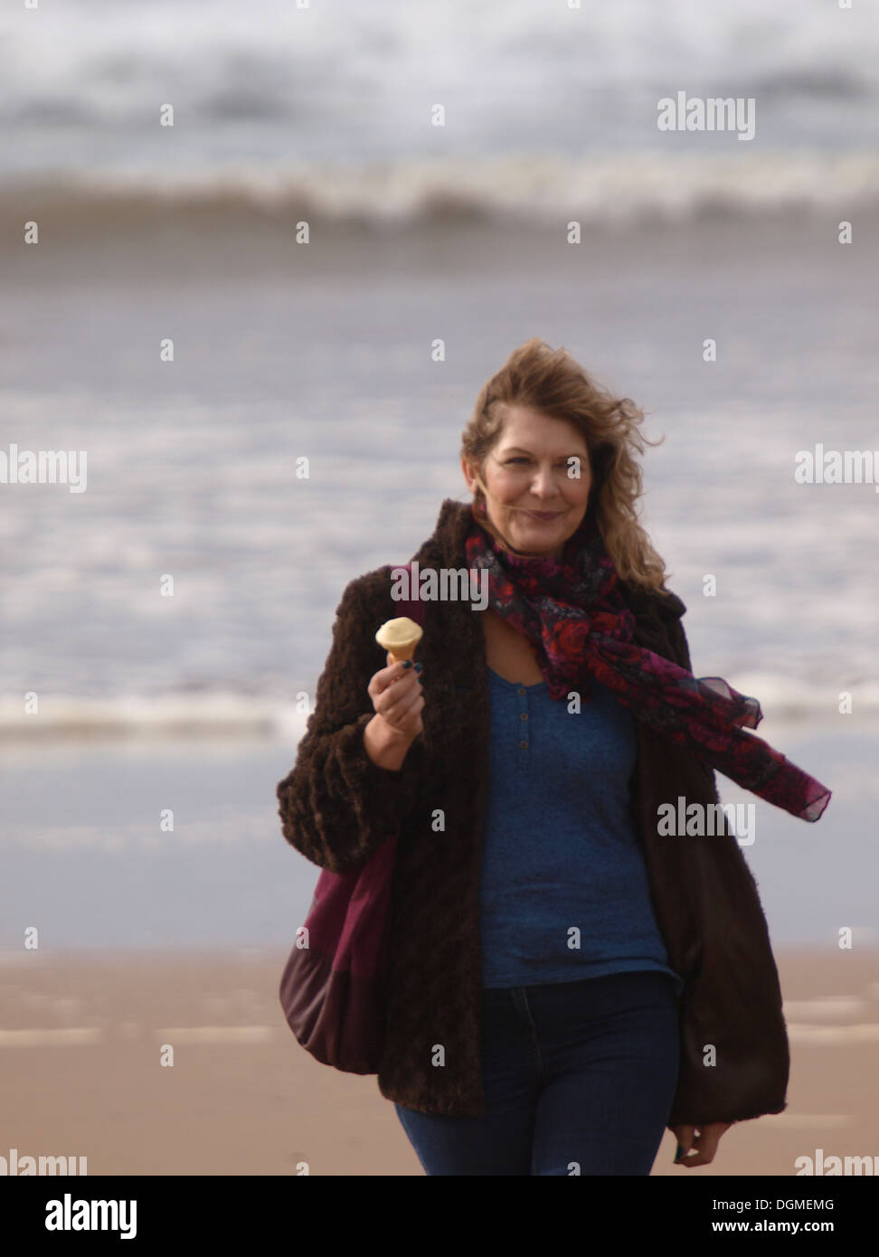 Middle aged woman walking on the beach with an ice-cream in winter, Bude, Cornwall, UK Stock Photo