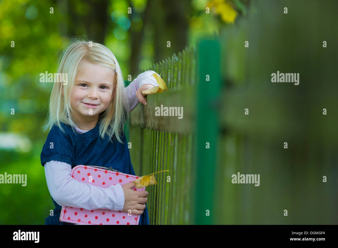Girl, 4 years old, holding a suitcase leaning against a wooden fence Stock Photo