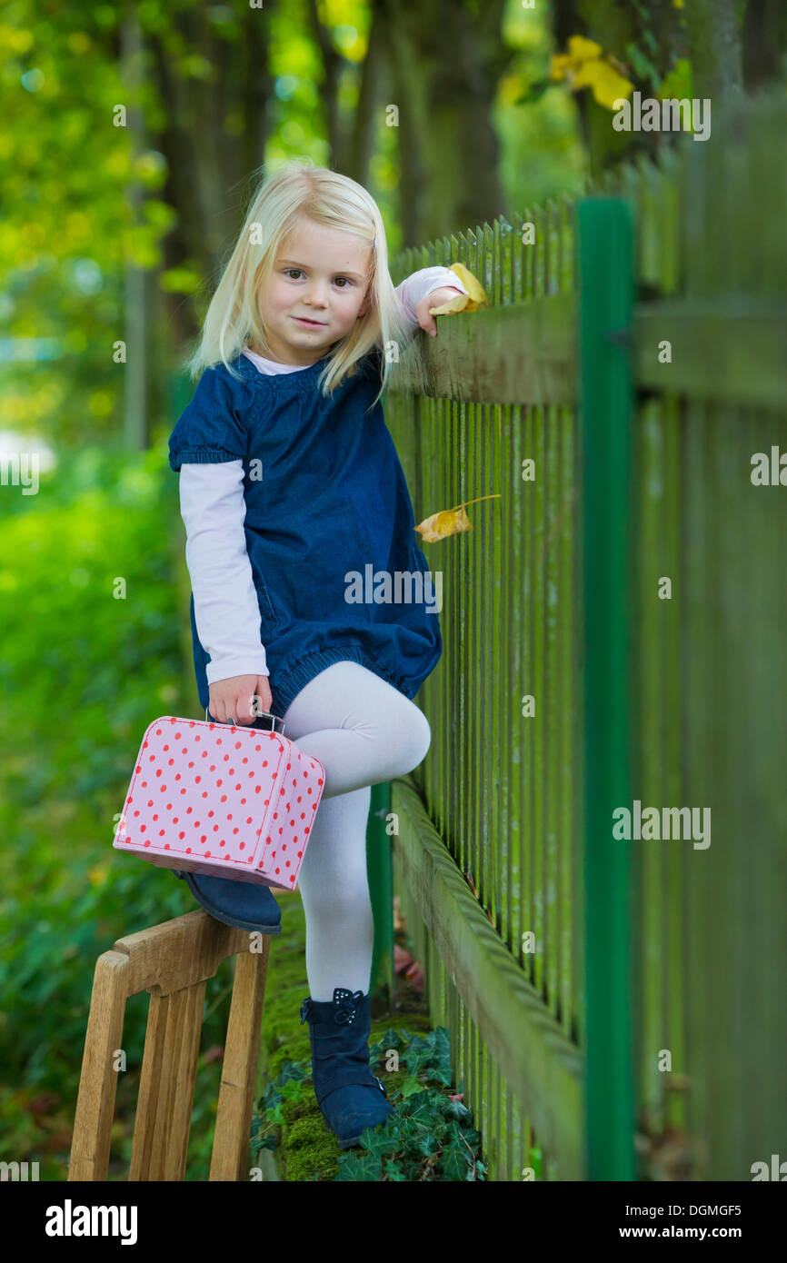 Girl, 4 years old, holding a suitcase leaning against a wooden fence Stock Photo