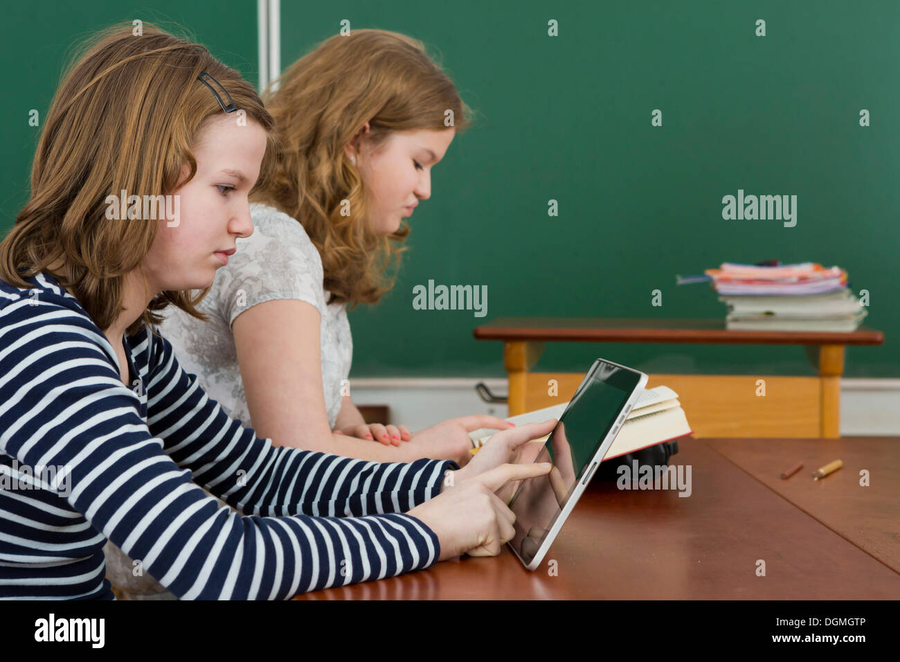 Two schoolgirls in the classroom, one is reading a book, the other is using a tablet PC, Germany Stock Photo
