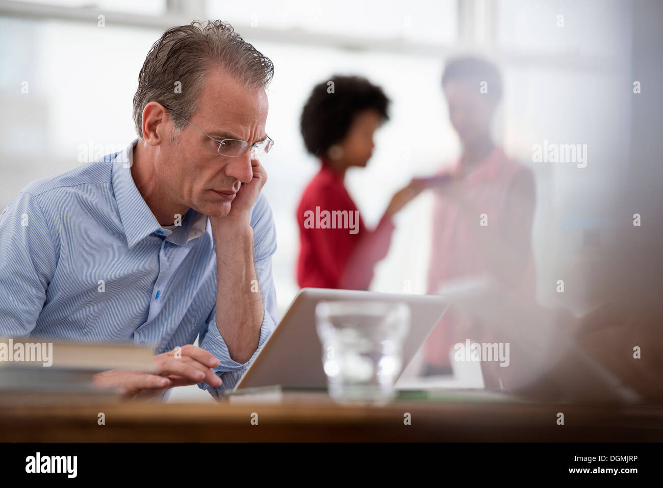 Office life. A man seated at a computer laptop leaning on one arm, and two women in the background. Stock Photo