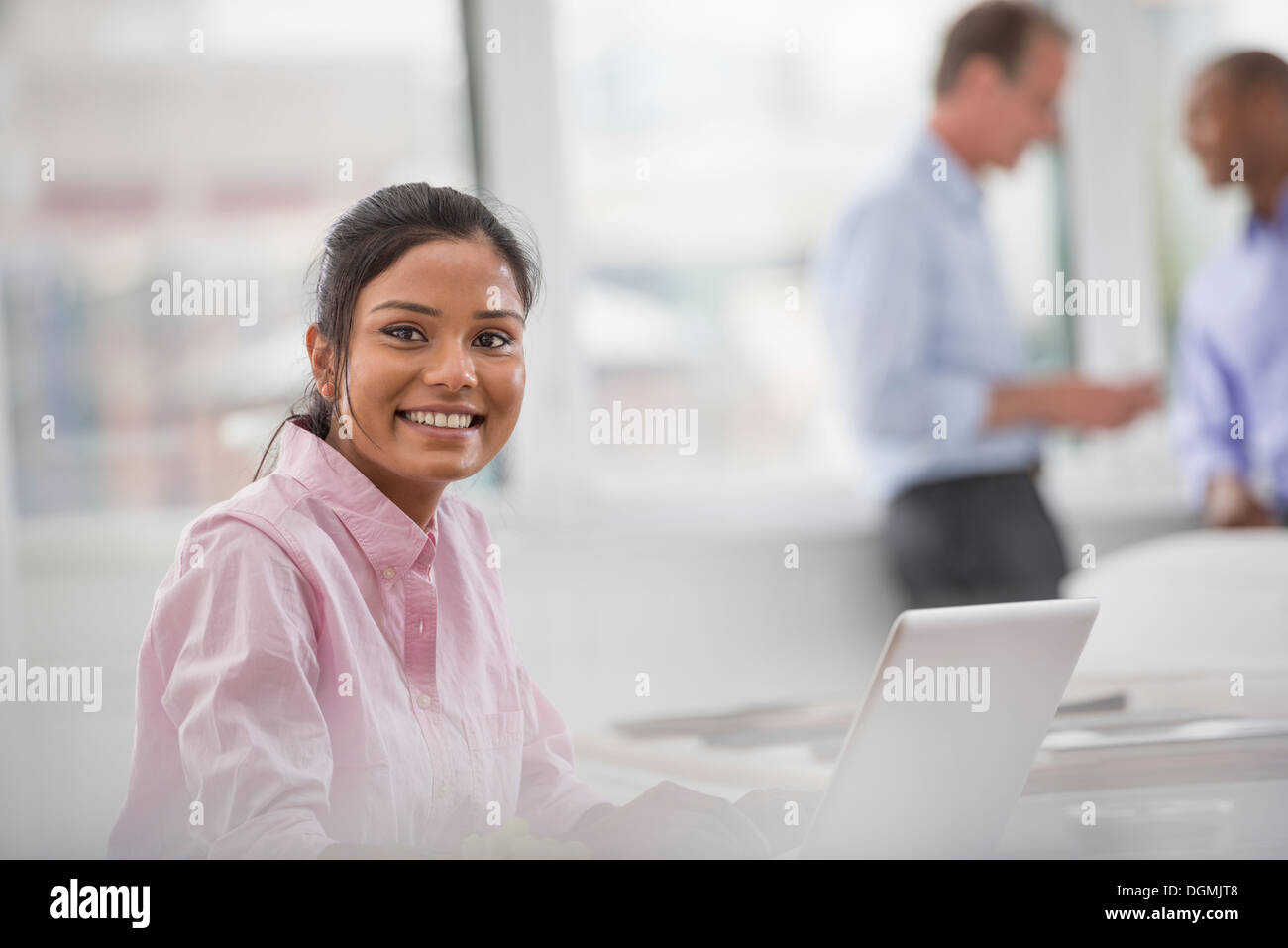 Office life. A woman sitting at a desk using a laptop computer. Two men in the background. Stock Photo