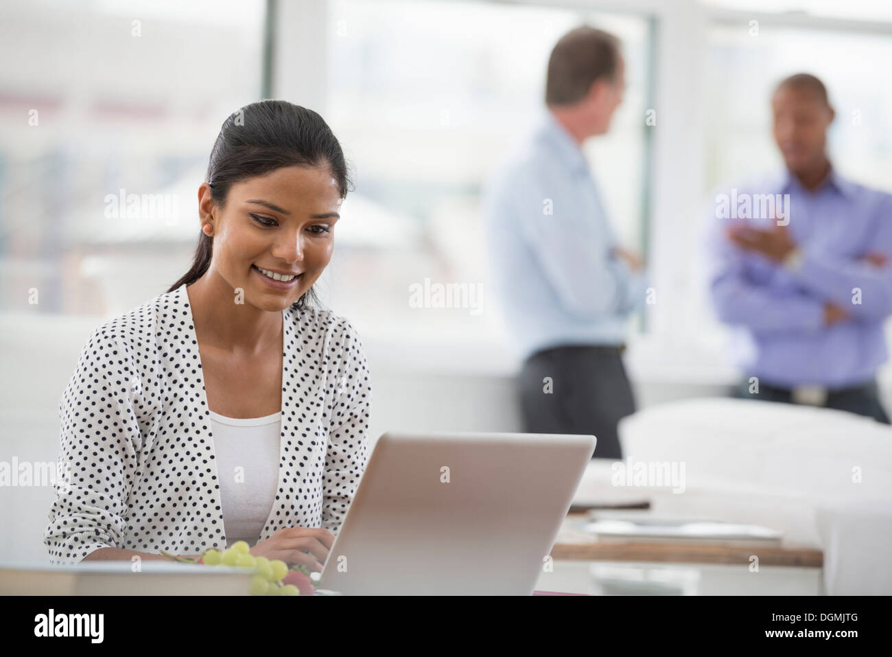 Office life. A woman sitting at a desk using a laptop computer. Two men in the background. Stock Photo