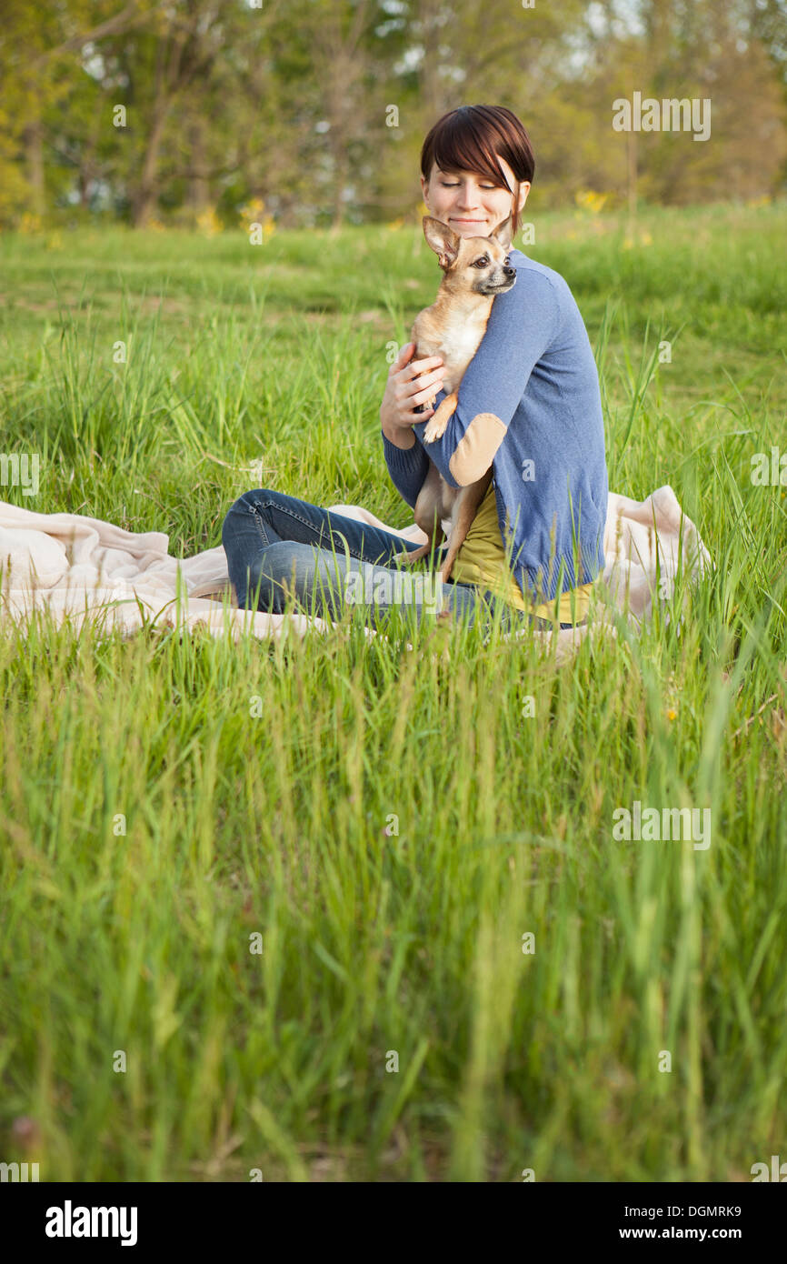 A young woman sitting in a field, on a blanket, holding a small chihuahua dog. Stock Photo