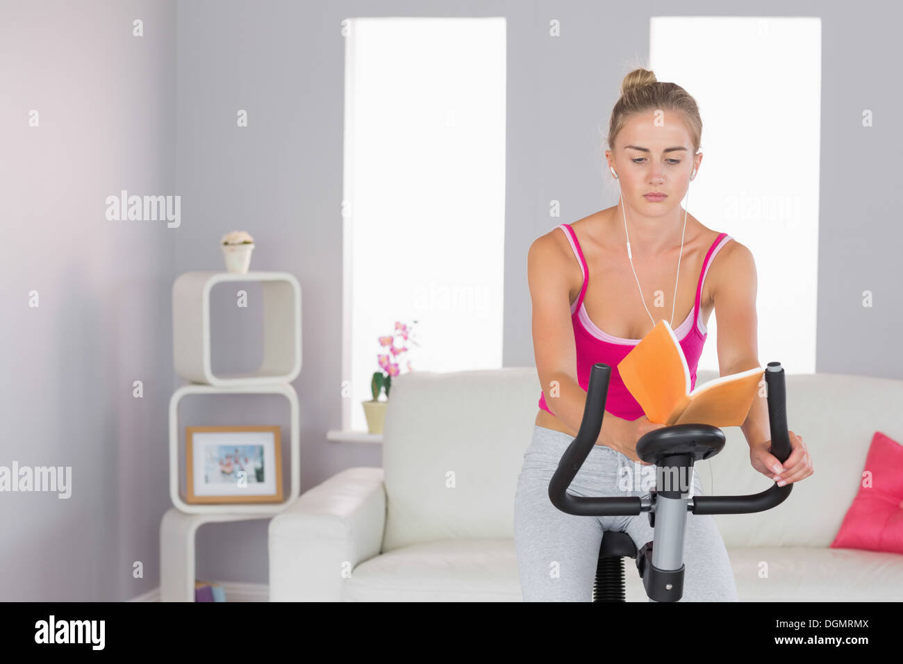 Sporty focused blonde training on exercise bike reading a book Stock