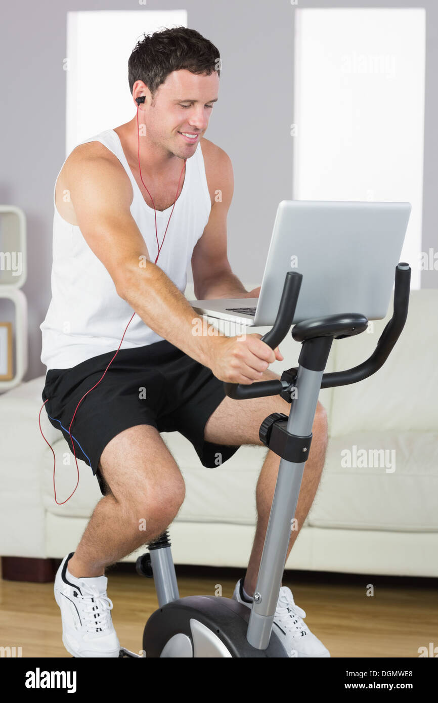 Sporty man with earphones exercising on bike looking at laptop Stock Photo