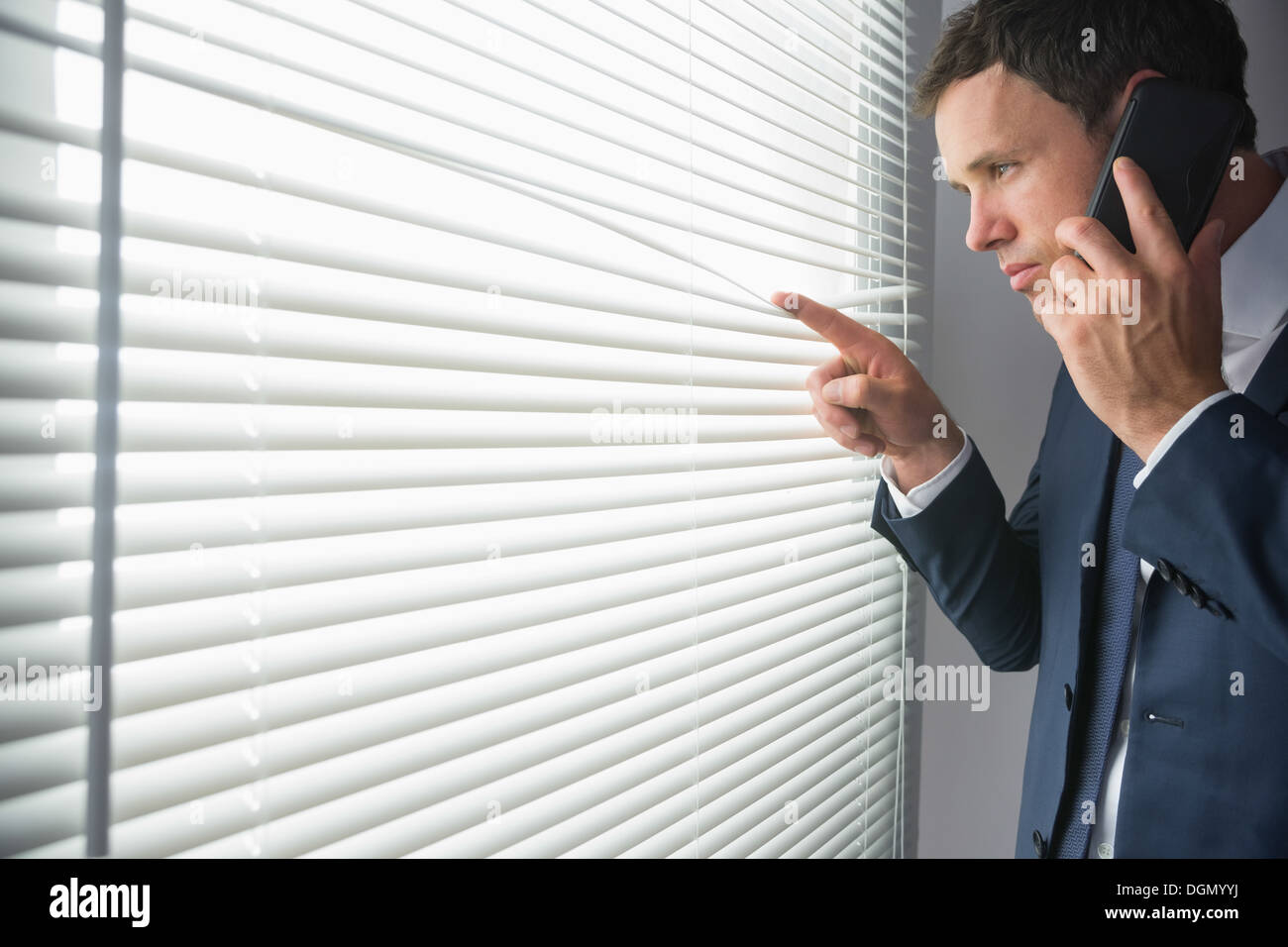 Unsmiling handsome businessman looking through roller blind phoning Stock Photo