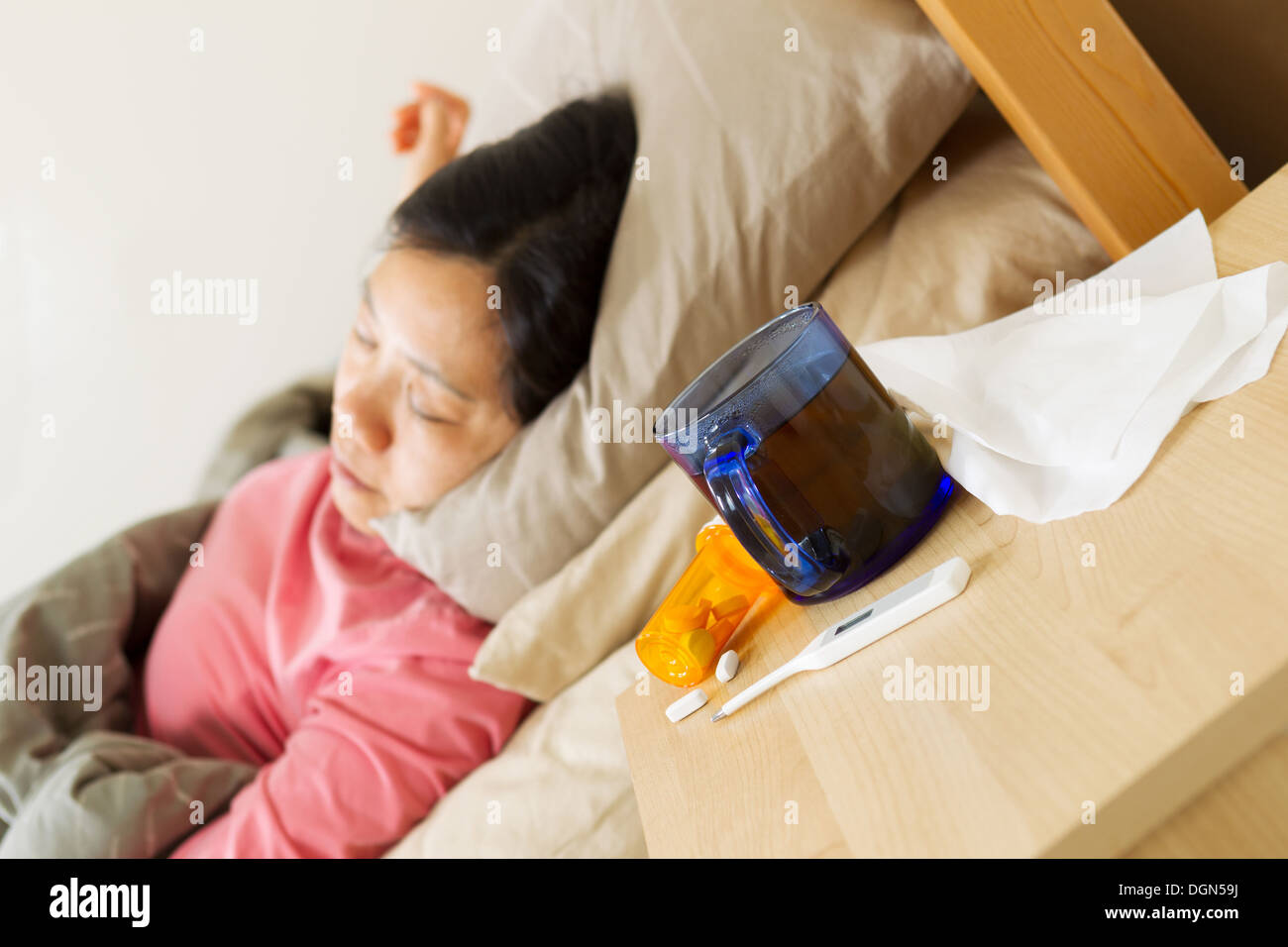 Horizontal photo of glass of hot tea, thermometer, tissues, and medicine on night stand with woman lying on her back while sick Stock Photo
