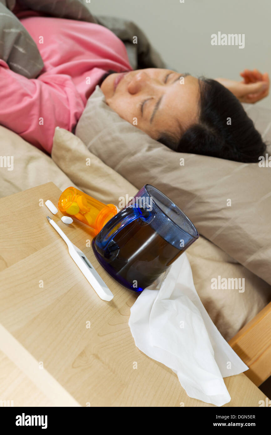 Vertical photo of glass of hot tea, thermometer, tissues, and medicine on night stand with woman