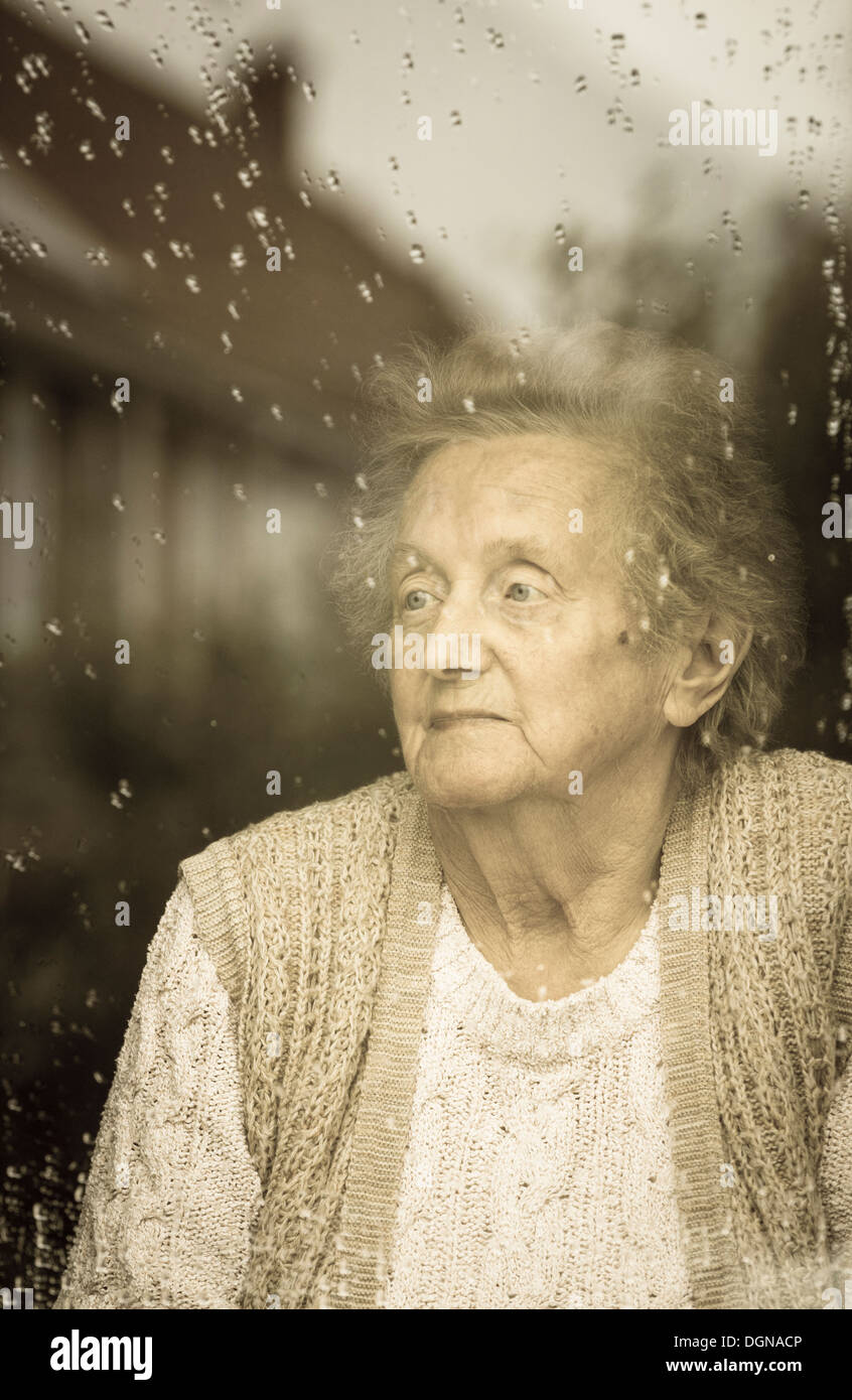 Ninety year old woman looking out of window on a rainy day. Self isolation, quarantine, Coronavirus, social distancing concept... Stock Photo