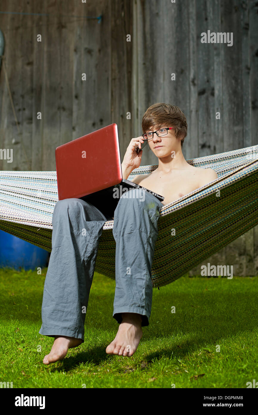 Teenager sitting in a hammock with a laptop speaking on his mobile phone Stock Photo