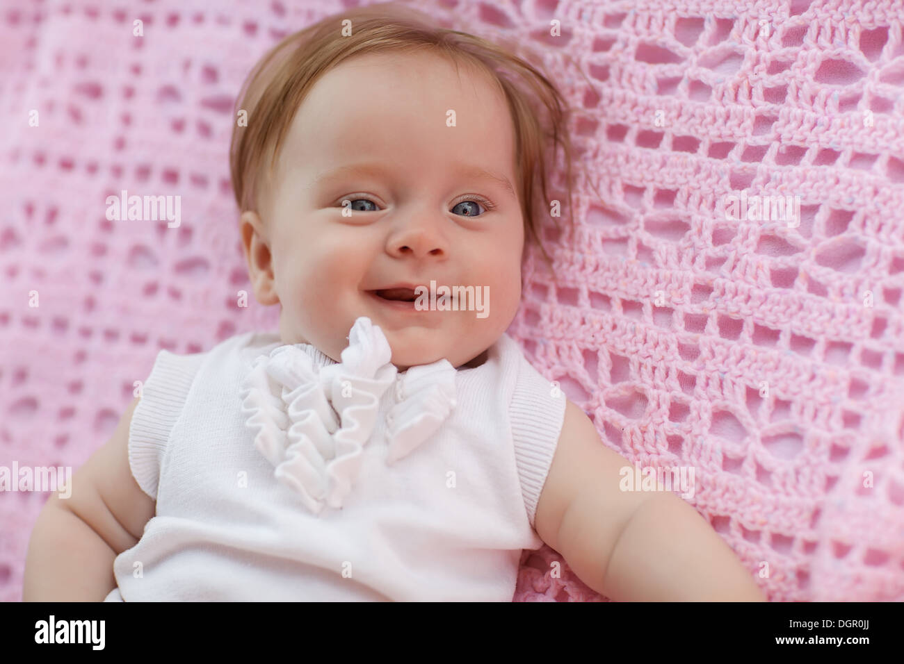 Sweet little baby girl lying on her back wearing a white jacket, looking at the camera and laughing back. Stock Photo