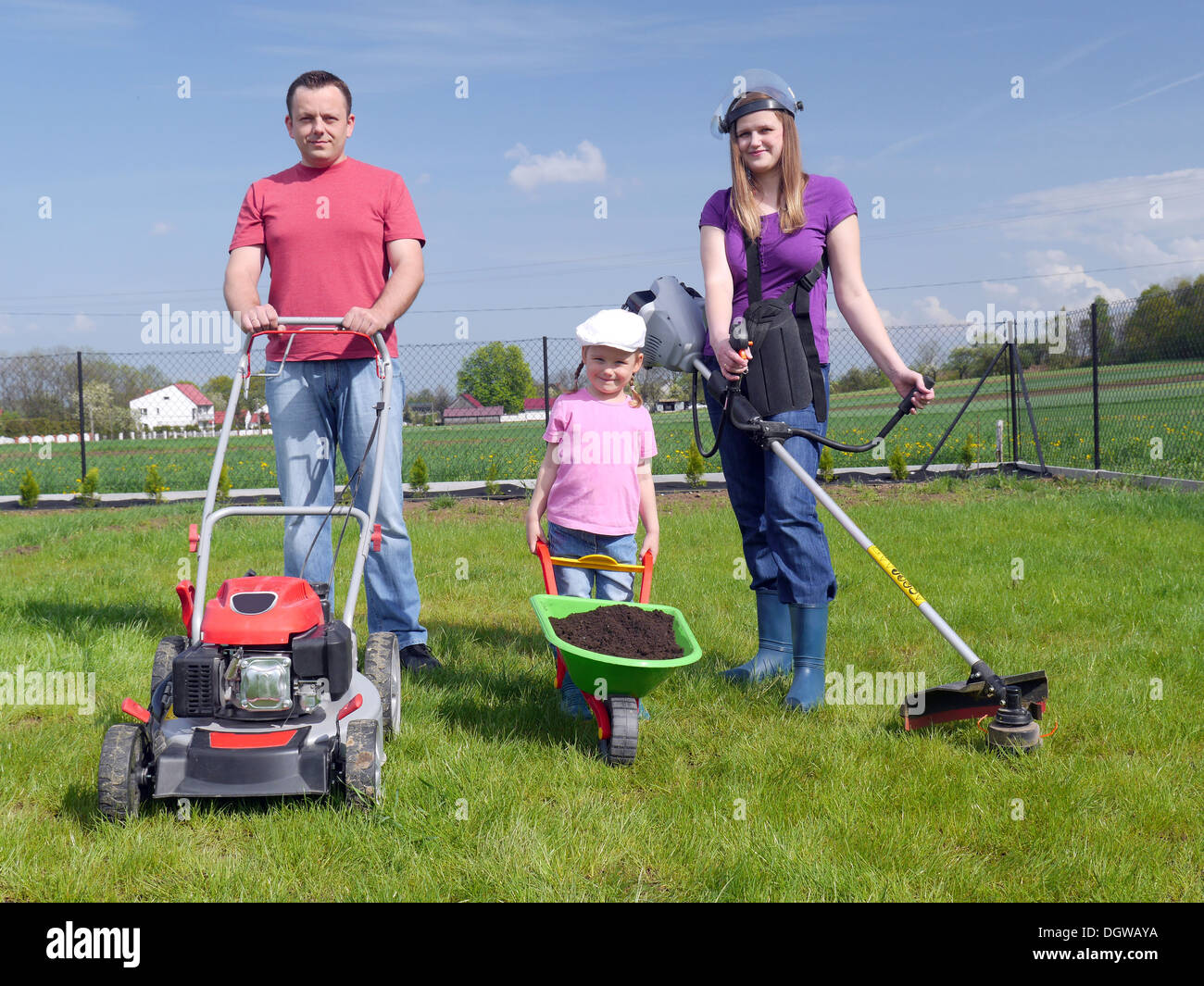 Dad standing with power mower, mom holding strimmer and their cute kid posing with green barrow full of soil Stock Photo