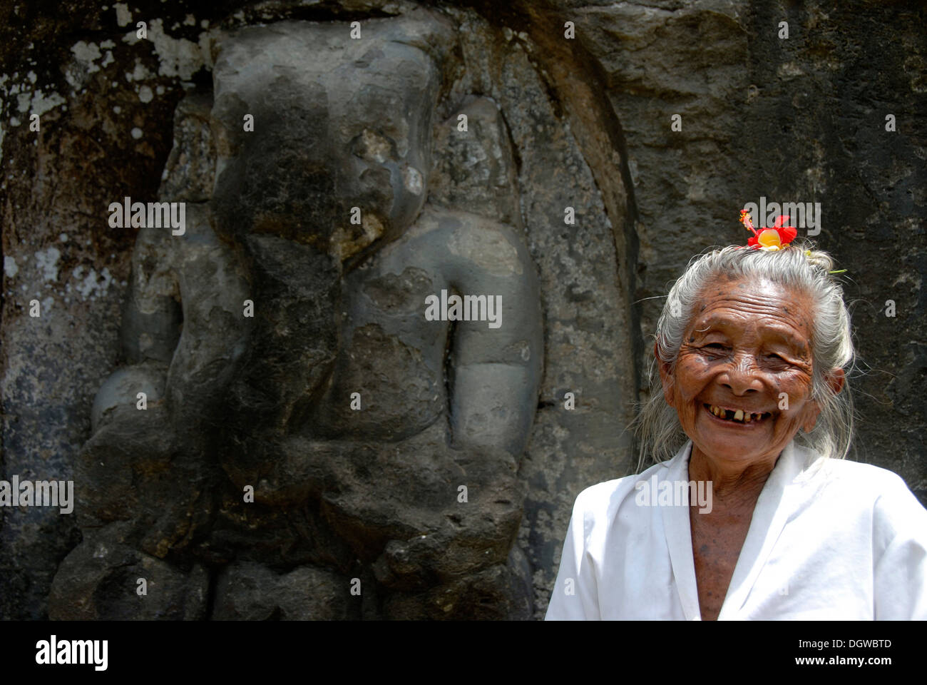 Bali Hinduism, portrait of an old Balinese woman with flower in her hear laughing in front of rock relief, God Ganesh or Ganesha Stock Photo