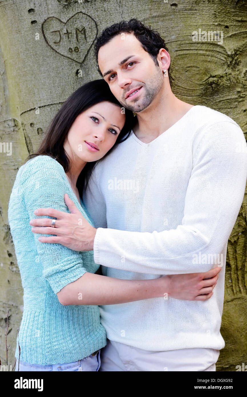 Young woman leaning on the shoulder of a young man in front of a tree with a carved heart, Austria Stock Photo