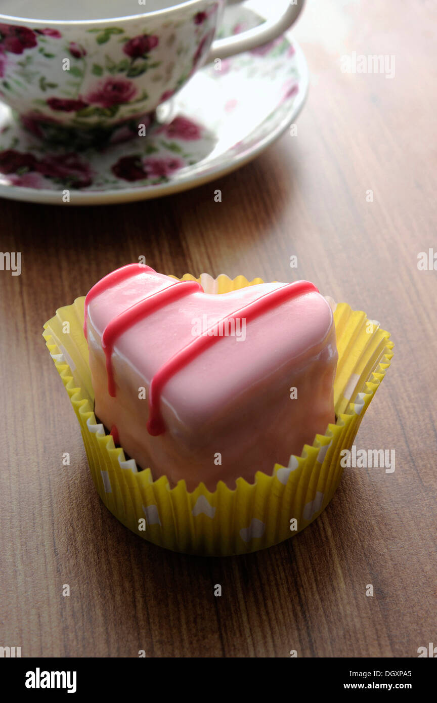 Pink heart-shaped petit four on a wooden surface beside a coffee cup Stock Photo