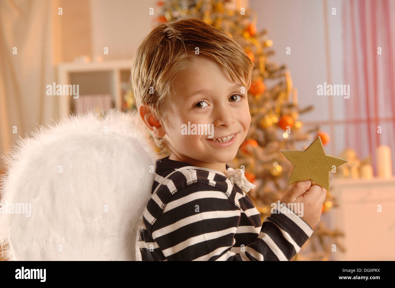 Boy wearing angel wings holding a golden star in front of a Christmas tree Stock Photo