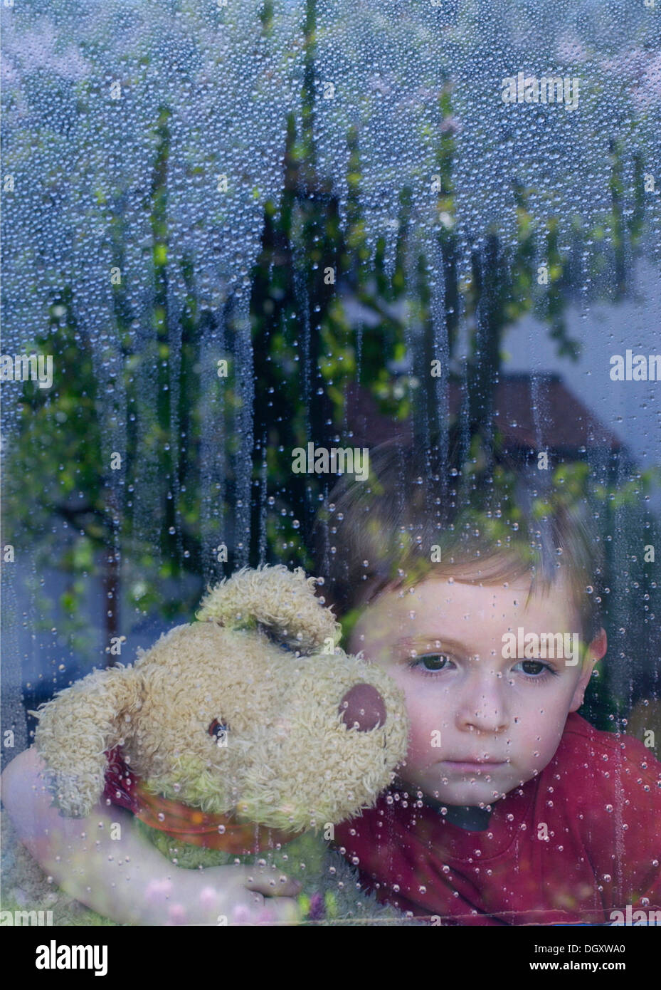 Boy looking sadly out of a window with water drops, holding a stuffed animal in his arms Stock Photo