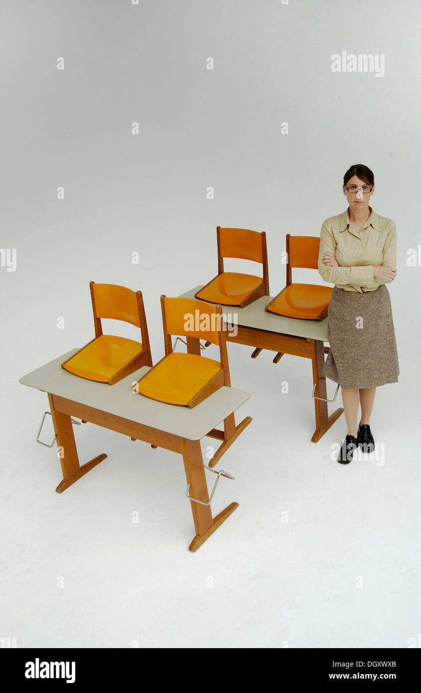 Strict female teacher standing next to old school desks with orange chairs Stock Photo