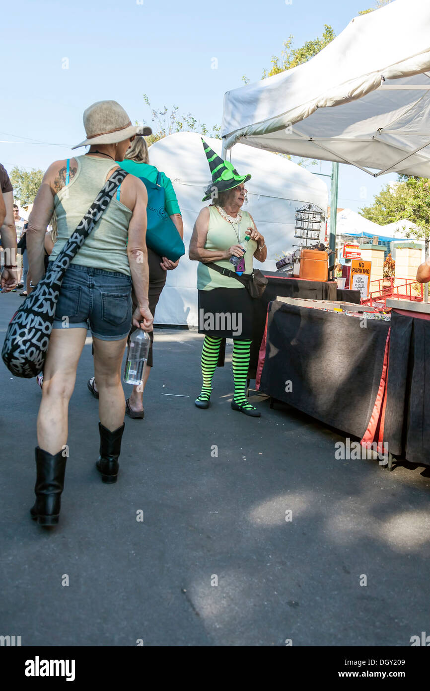 Craft vendor wearing black and neon green striped witch hat and tights white woman wearing boots, shorts and hat walks by. Stock Photo