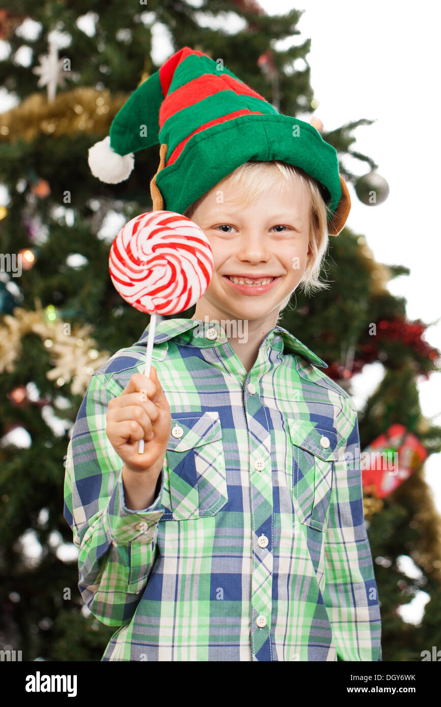 A cute boy dressed as Santa's helper or an elf is holding a red lollipop and smiling in front of a Christmas tree. Isolated on w Stock Photo