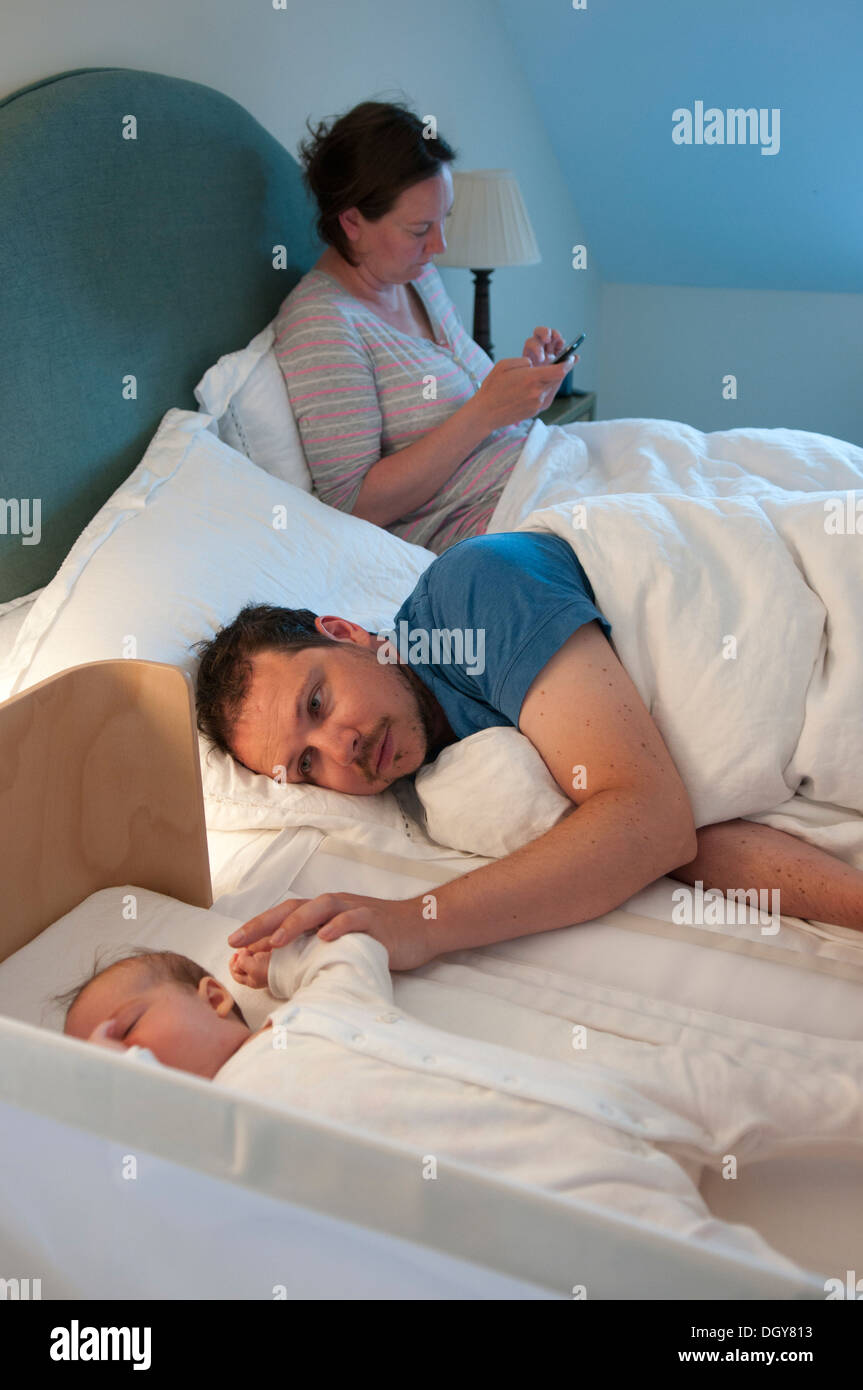 New parents in bed, dad comforting baby in crib and mum using mobile phone Stock Photo