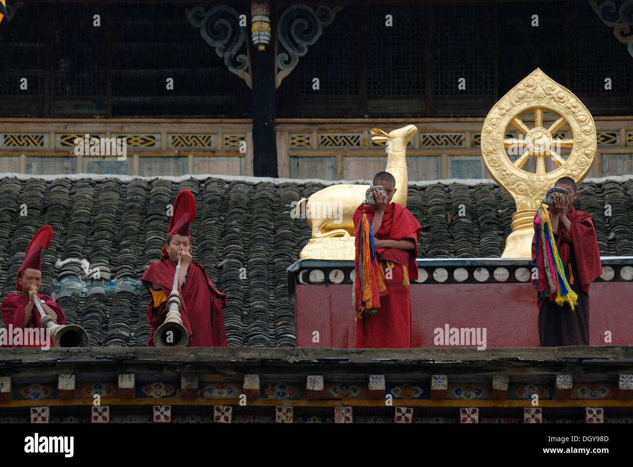 Monks with red robes and caps calling to prayer with conch horns and monastery trumpet on the roof balcony of the Lhagang Stock Photo