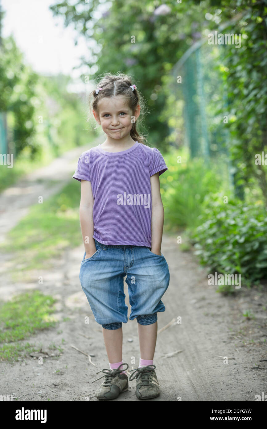A young smiling girl with hands in her pockets, standing on a dirt road Stock Photo
