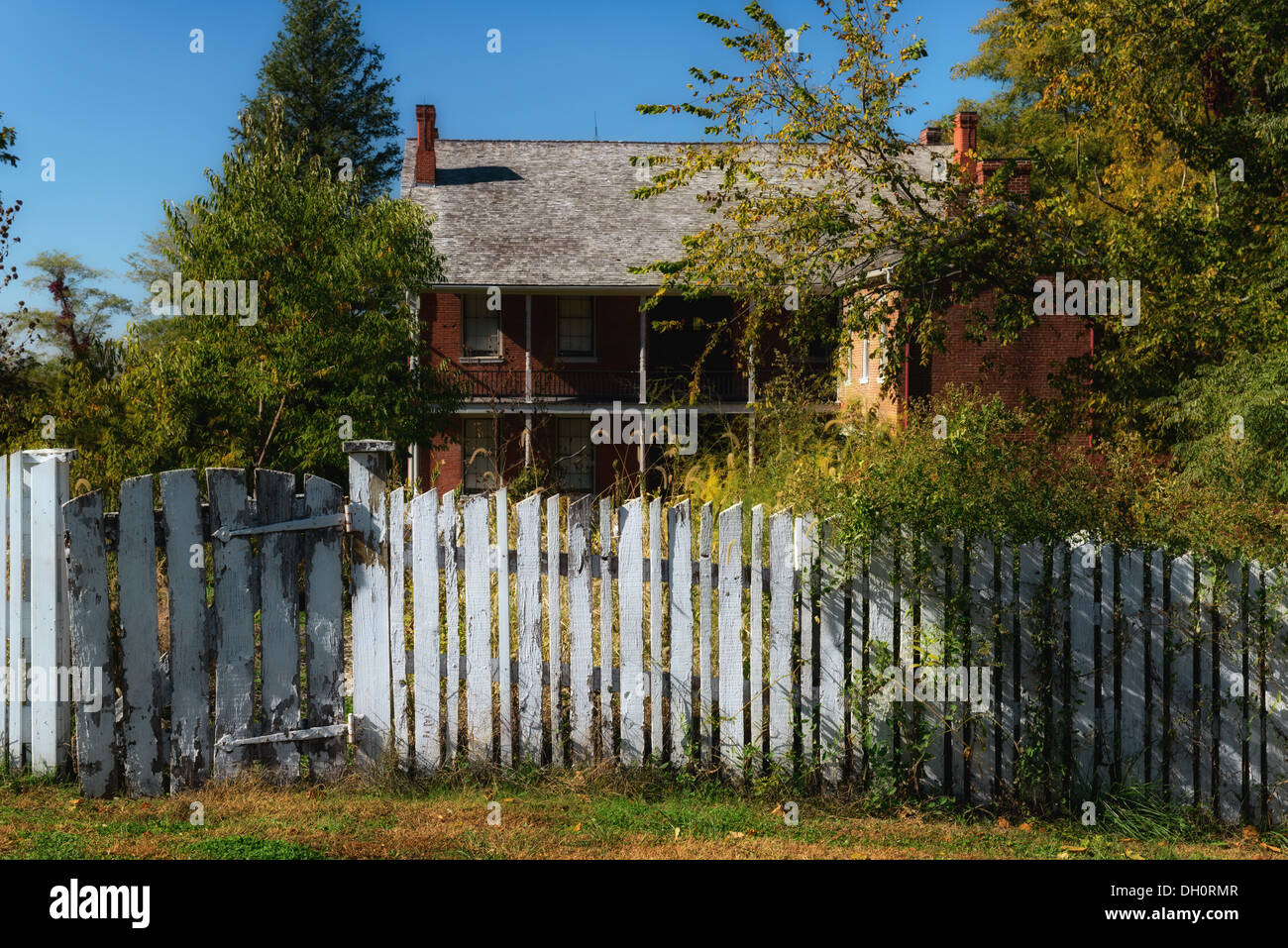 Photograph of a two story brick house surrounded by an unkempt yard and an old white picket fence Stock Photo