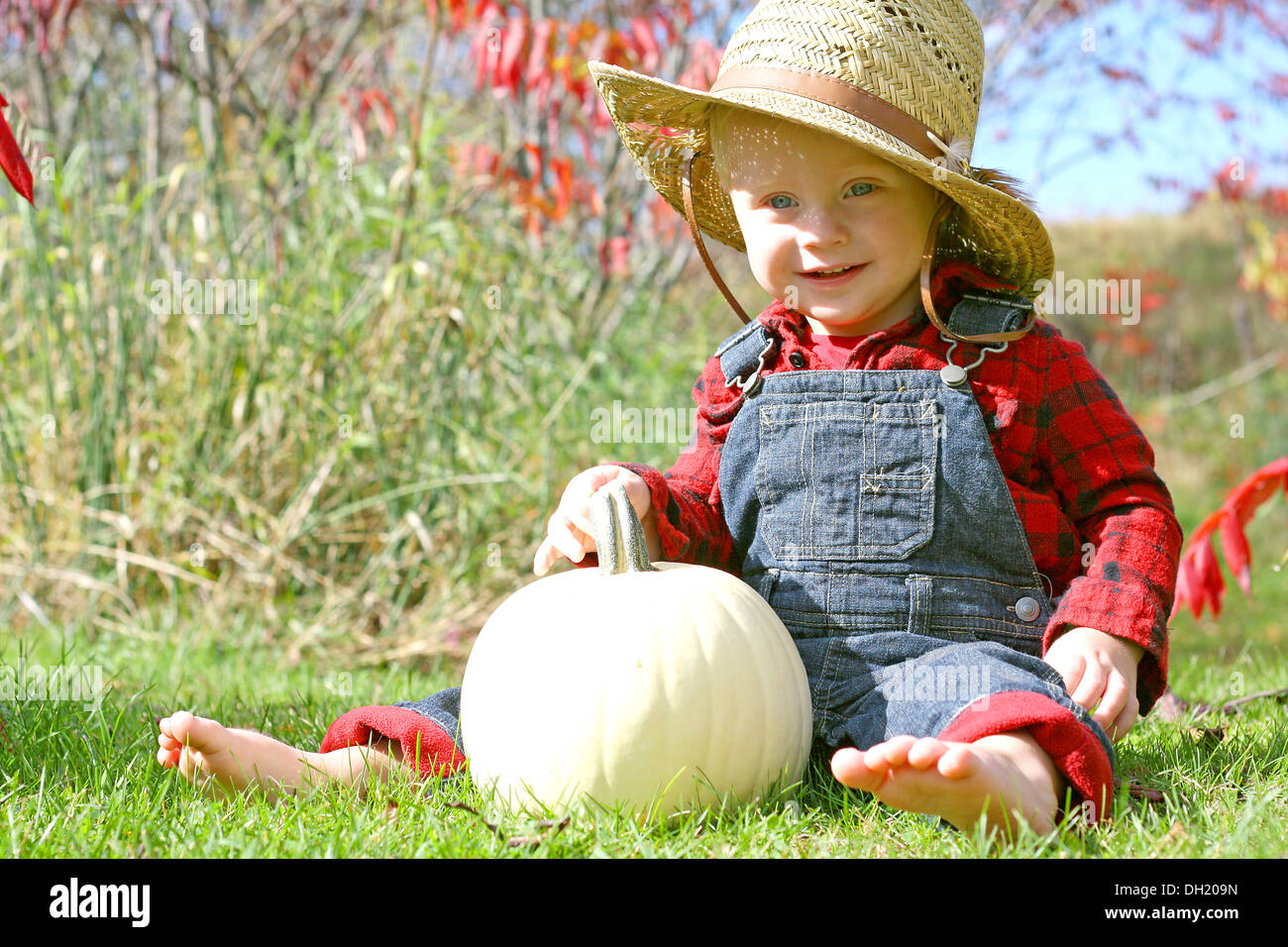 a cute, smiling baby boy is sitting outside in the grass with a white pumpkin on a sunny autumn day, dressed like a farmer Stock Photo