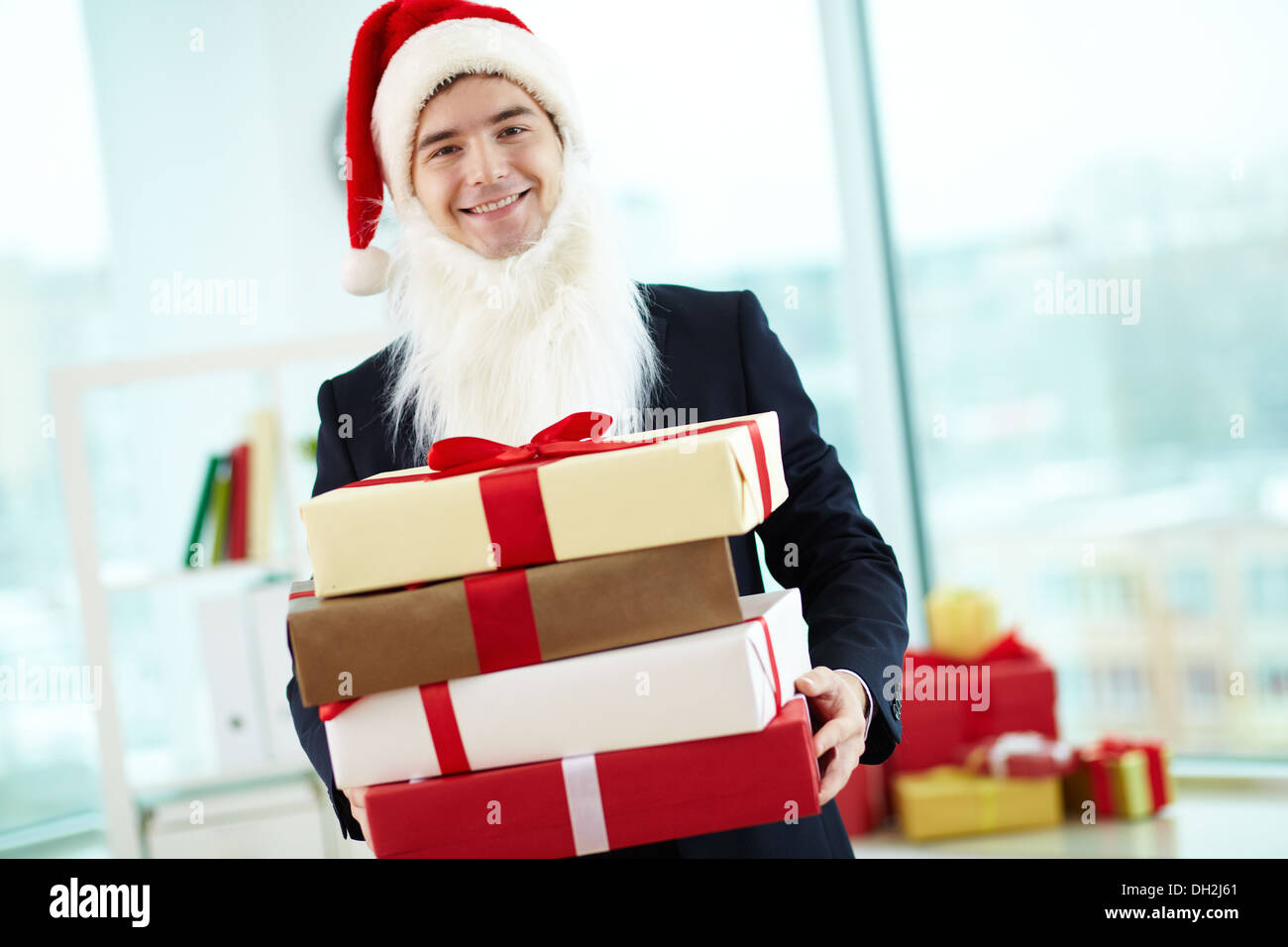 Image of happy businessman in Santa cap and beard holding stack of gifts and looking at camera in office Stock Photo