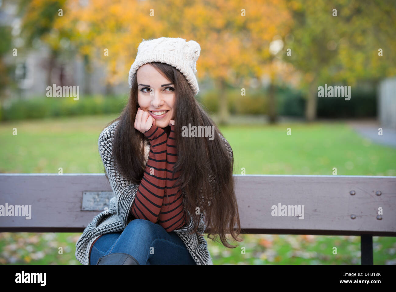 Young woman in a park during autumn sitting on a bench wearing a woolly ...