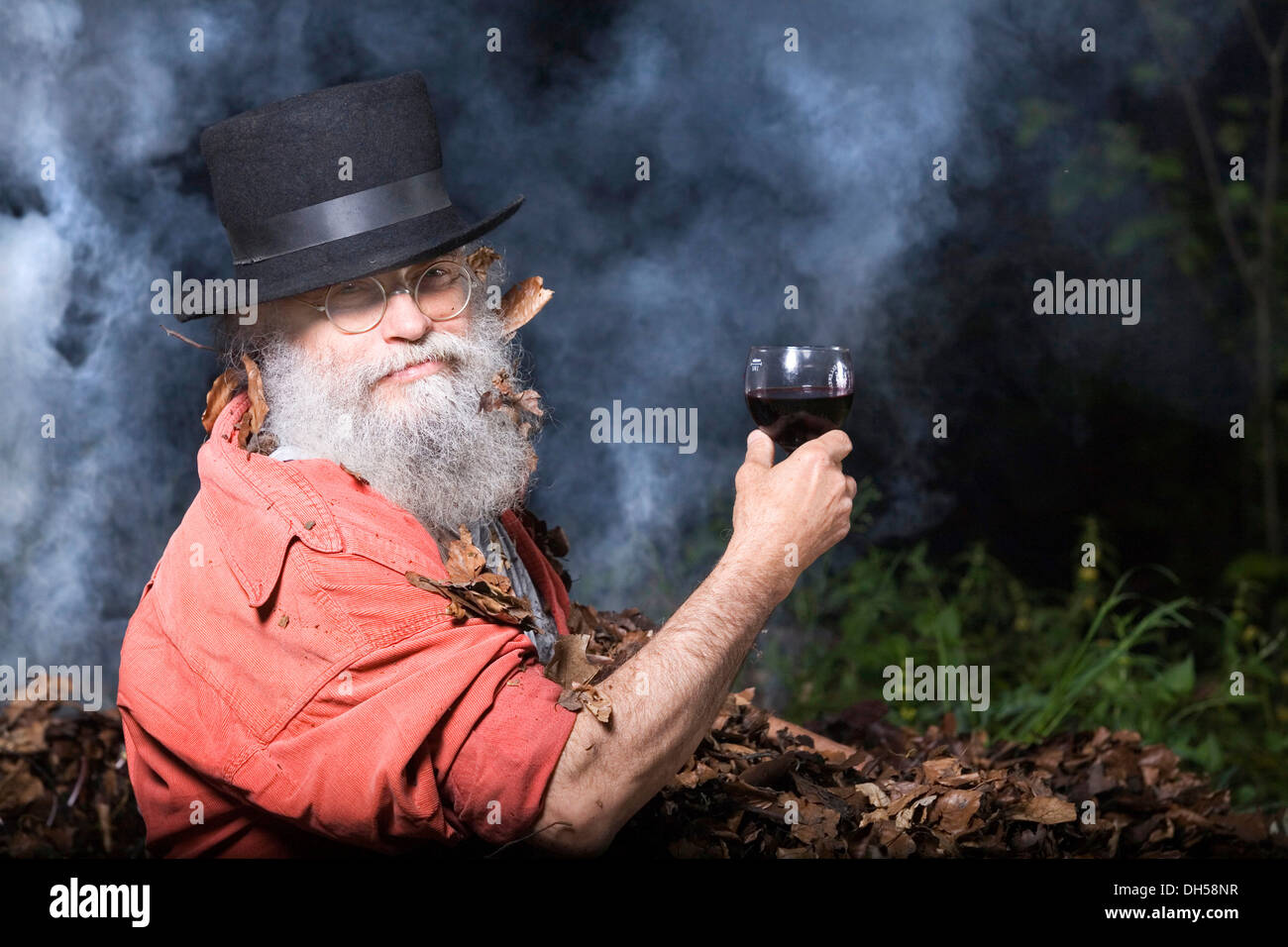 Elderly man with a long beard wearing a top hat and glasses, lying in a pile of autumn leaves and holding a glass of red wine Stock Photo