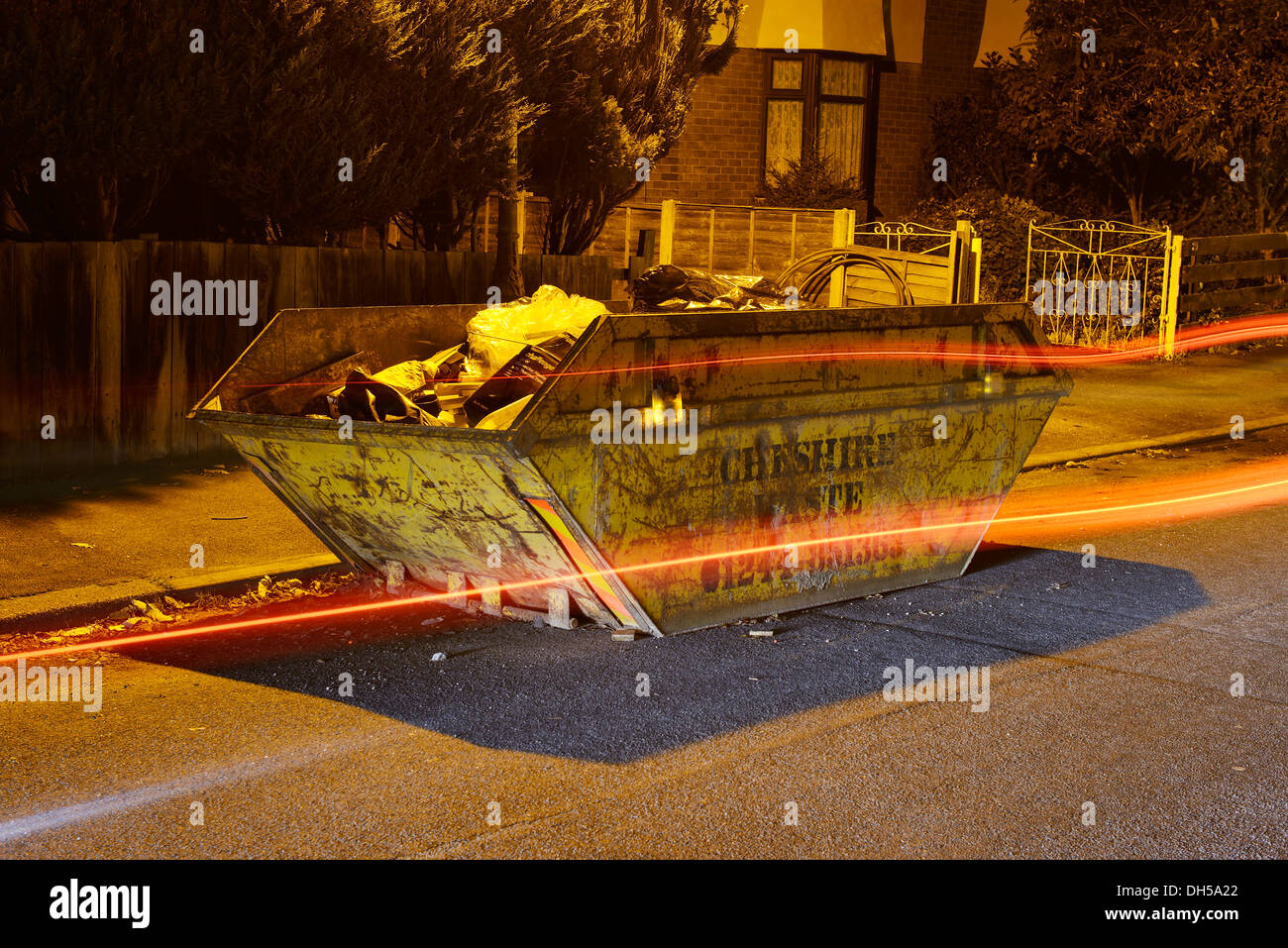 Large metal skip positioned on the side of a road as traffic drives past Stock Photo