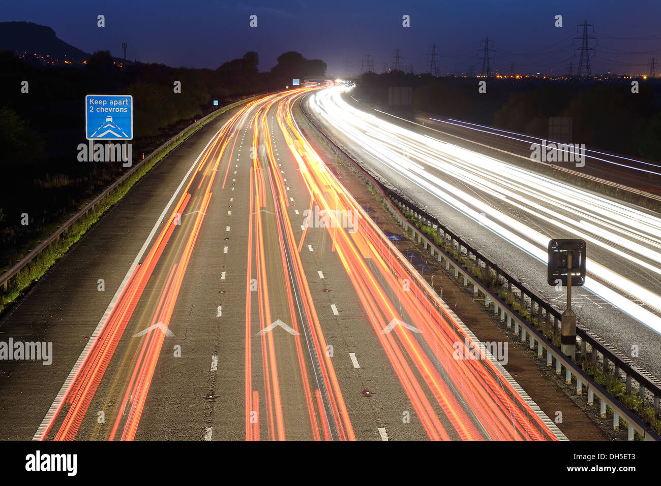 Long time exposure of evening traffic on the M56 motorway showing a Keep Two Chevrons Apart sign UK Stock Photo