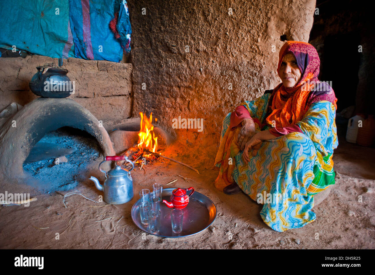 Elderly Berber woman preparing tea while squatting on the ground in front of a clay oven and a small fire, Erg Chebbi Stock Photo