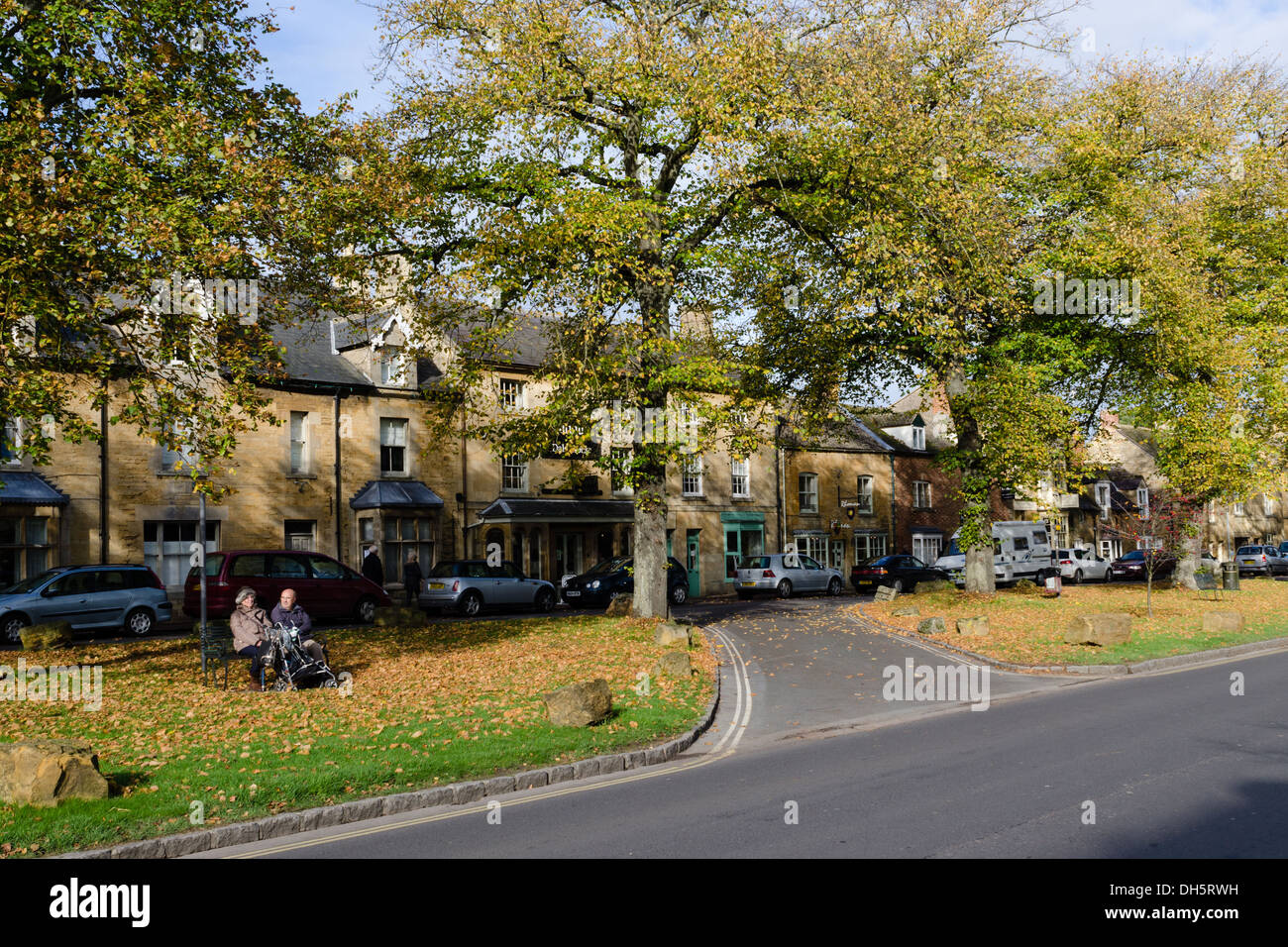 Couple sitting on a bench in front of a row of shops Moreton-in-Marsh High Street in the Cotswolds Stock Photo