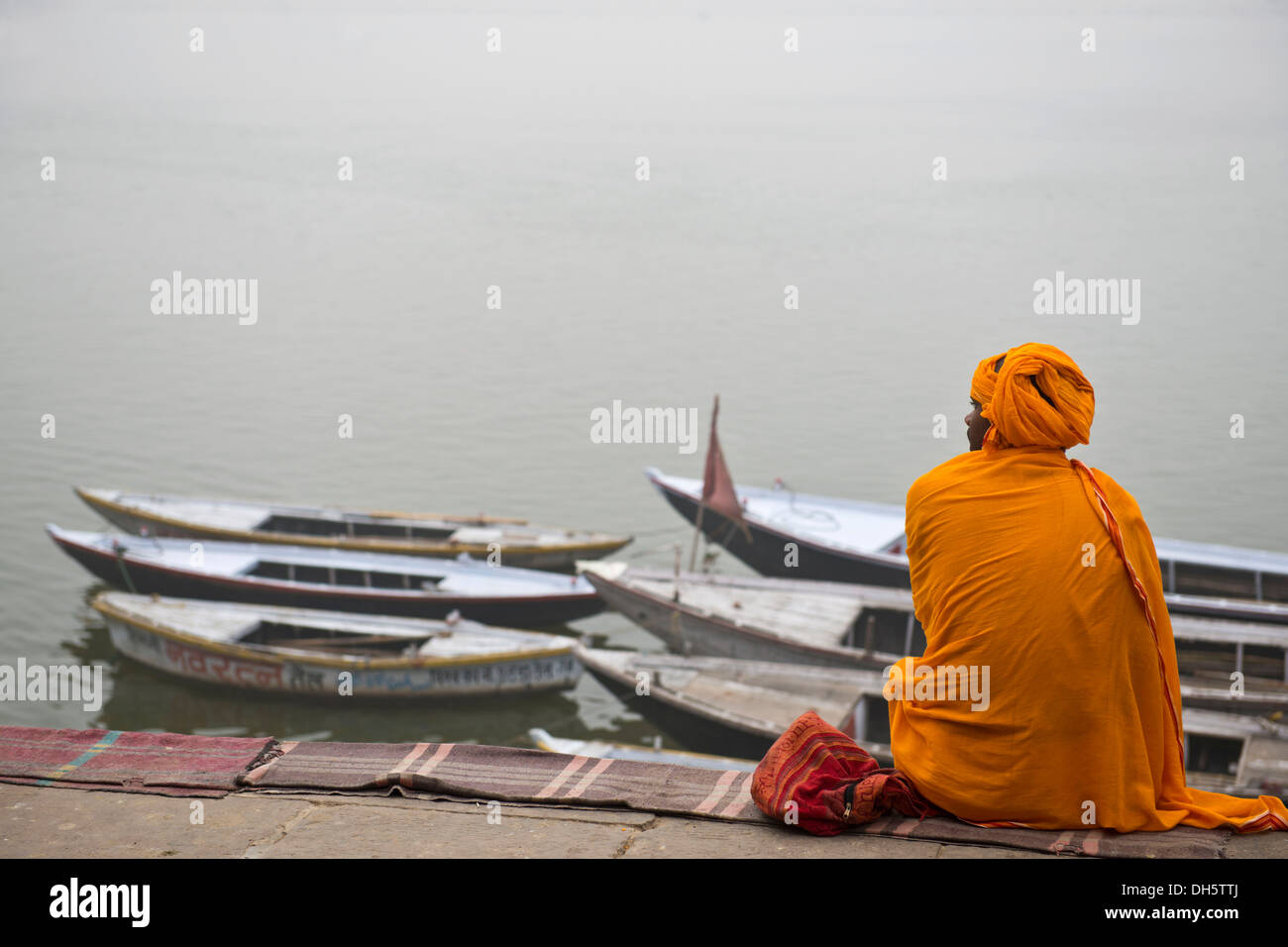 Brahmin or Hindu priest sitting on a blanket on the banks of the Ganges River with several boats tied to the shore, Varanasi Stock Photo