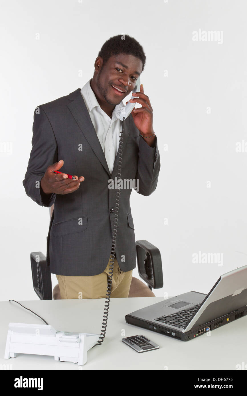Young dark-skinned man standing at a laptop, holding a phone and smiling Stock Photo