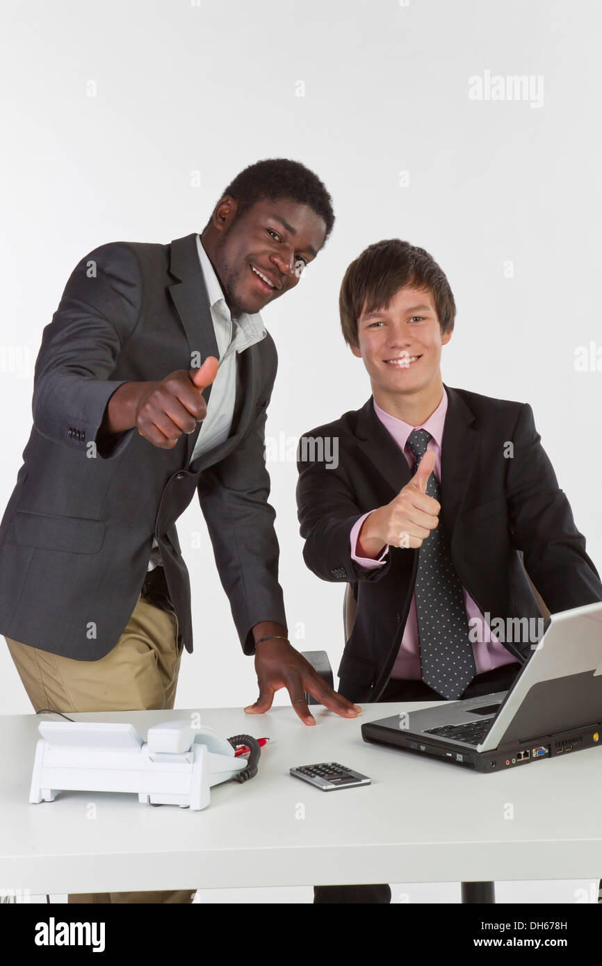 Two young businessmen of different races with a laptop and making a thumbs-up gesture Stock Photo